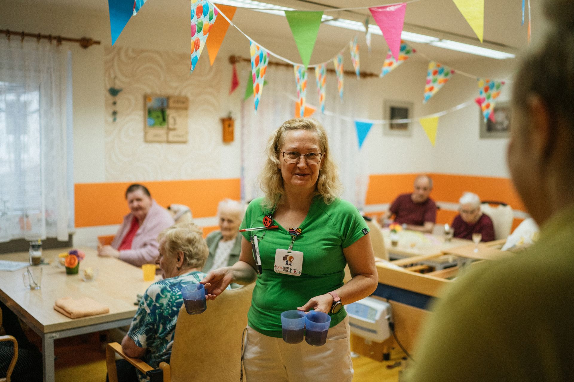 A smiling caregiver serves drinks to residents in a decorated group home common room